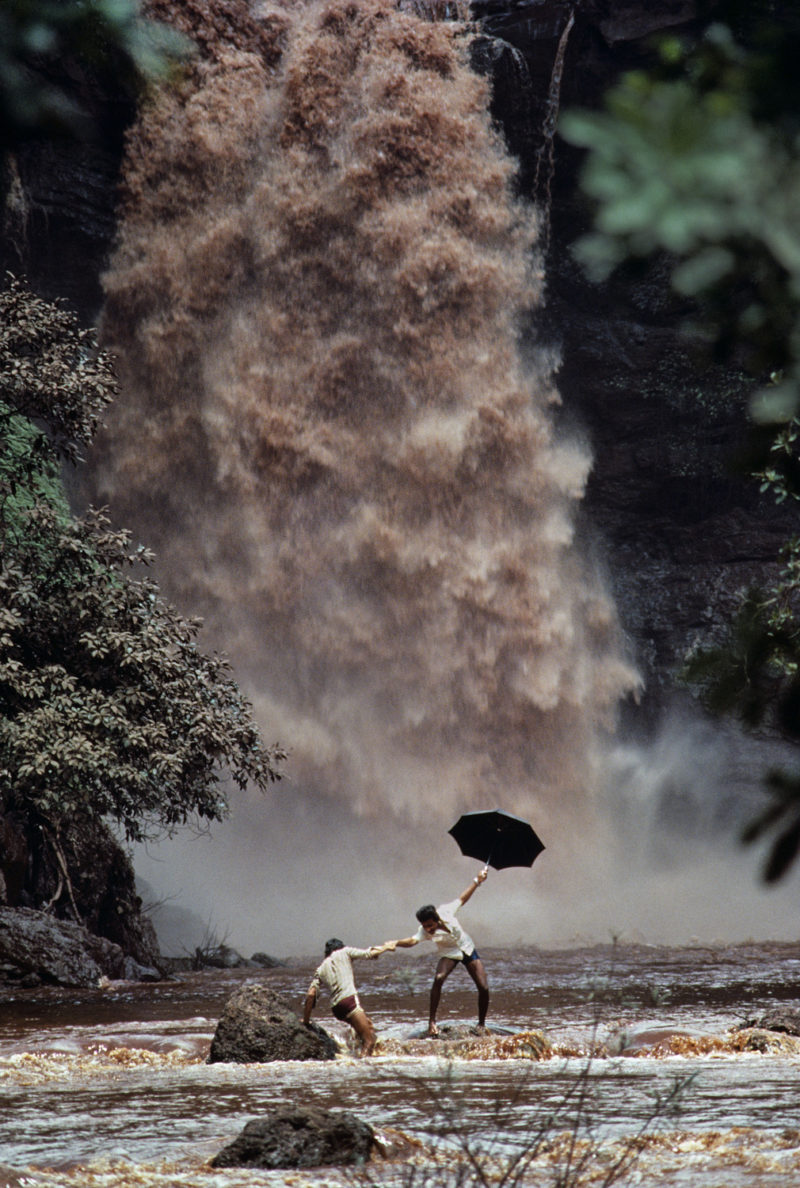 Steve McCurry - Two men crossing a swollen river after the bridge was washed away, Goa, India