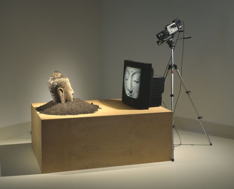 Nam June Paik – Buddha Watching TV, 1974/1997, Stone sculpture, soil, closed-circuit video camera, video monitor, tripod, plywood base, Virginia Museum of Fine Arts