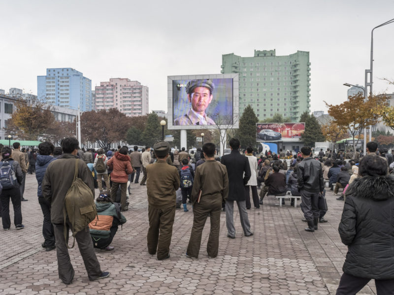 Carl De Keyzer - Pyongyang Station, Pyongyang. Locals and travelers watch a film on a public screen. This screen in the center of the city regularly shows movies throughout the day. It is also used for important news broadcasts, such as the announcement of nuclear or missile tests, for example. 9 November 2015 11:00 AM