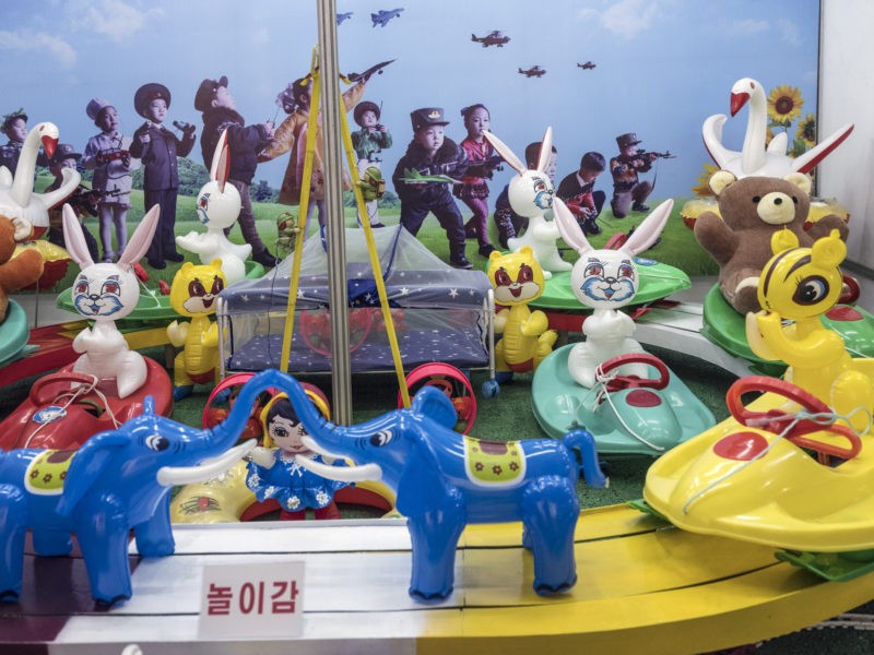 Carl De Keyzer - Toys, with a background image depicting children dressed as soldiers and doctors, playing with remote-control vehicles. Three Revolutions Exhibition. Pyongyang. 29 May 2017 4:00 PM