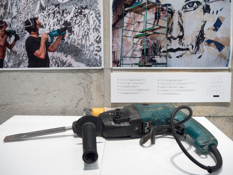 Hammer drill used by street artist Vhils alongside photos of him creating an artwork by Martha Cooper, installation view, Tools of the Trade, Hong Kong, 2021